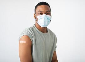Coronavirus Vaccination. Portrait Of Vaccinated African Guy Wearing Protective Face Mask Looking At Camera Posing With Adhesive Tape On Arm After Vaccine Injection Over White Background photo