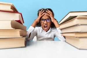 School Overload Concept. Afro Schoolgirl Screaming Clutching Head Sitting Between Book Stacks. Yellow Background, Studio Shot photo