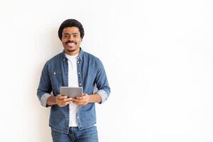 African American man is standing while holding a tablet in his hands. He appears focused on the screen, possibly reading or watching something. The background is white with copy space photo