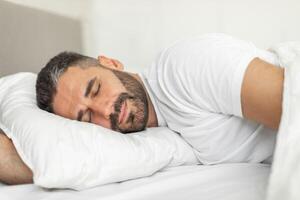 A man with a beard is lying on his side in a bed, sleeping soundly. His eyes are closed, and his face has a relaxed expression. He is wearing a white t-shirt and is covered with a white blanket. photo