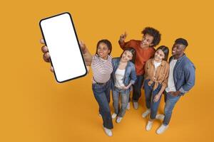 A group of five diverse friends stand together on a yellow background. The person on the left is holding a smartphone with a blank screen. All of the friends are smiling and looking at the camera photo