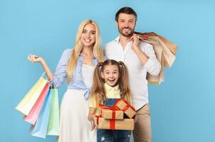 A happy family, a father, mother, and daughter, are standing together against a light blue background. The mother is holding a stack of colorful shopping bags in her left hand photo