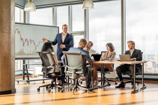 A businessman stands in front of a large screen displaying financial data while presenting to a group of colleagues sitting around a table in a bright and modern office. photo