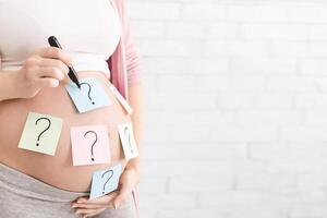 Concept of choosing baby name. Pregnant woman with question marks on paper stickers on tummy, panorama with empty space photo
