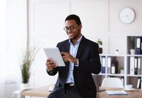 Modern technologies in business. Happy african american young businessman in suit using digital tablet in office, working online, reading marketing report, getting ready for presentation, copy space photo