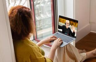 A woman sits by a window with a laptop in her lap, engaged in a call with a man who appears on the screen. She is typing on the keyboard with one hand while she looks at the screen photo