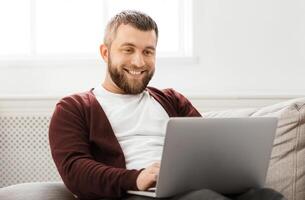 A man is seated on a comfortable couch, focused on a laptop screen in front of him. He appears engaged, possibly working or browsing. The room is well-lit, with a casual and relaxed atmosphere. photo