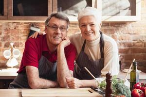 A senior couple, wearing aprons, are happily cooking together in their kitchen, smiling at each other, seemingly enjoying their time together. Ingredients and utensils are visible on the counter photo