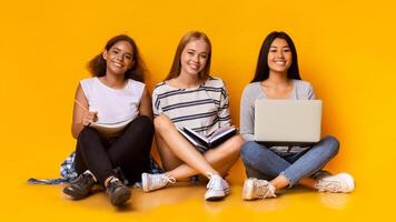 Three young cheerful multinational girls studying together with laptop, sitting on floor over yellow background photo