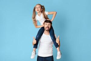 A father and daughter are playfully posing against a light blue background. The girl is sitting on her father's shoulders with her arms around his neck. photo