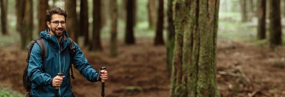 A man with a backpack and hiking poles walks through a forest trail on a cloudy day. He is smiling as he looks ahead, enjoying the peace of nature, copy space photo