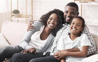 Happy family portrait. Smiling black mother, father and daughter cuddling and posing to camera on couch at home photo