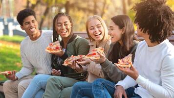 Hungry laughing international teenagers enjoying pizza while spending time in park photo