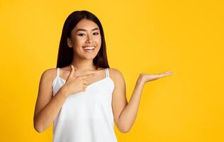 Asian smiling woman standing against a vibrant yellow background. She is pointing with her left hand and presenting with her right hand, creating a space for product placement or a visual message. photo