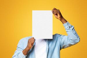 African american man covering his face with white blank paper, standing over yellow background in studio, copy space photo