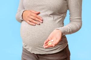 Unrecognizable Pregnant Girl Showing Pills In Hand Touching Belly Standing Over Gray Studio Background. Cropped photo