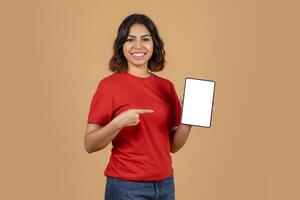 A young middle eastern woman with a bright smile is holding a tablet with a blank white screen. She is wearing a red t-shirt and blue jeans. The background is a light brown color. photo