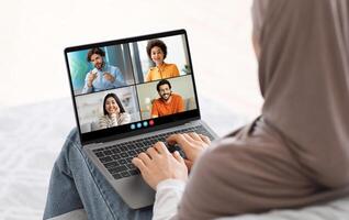 Muslim woman sits on a bed with her back to camera, using laptop to chat with three friends. She is wearing hijab and is typing on keyboard, actively participating in online conversation. photo