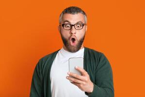 A bearded man with glasses is expressing shock as he looks at his smartphone. He is dressed casually in a green sweater and a white t-shirt. The background is a solid orange color photo