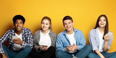 A cheerful quartet of young friends are seated side by side, casually dressed, each holding a smartphone, amused and engaged in a moment of digital interaction, against a vibrant yellow backdrop photo
