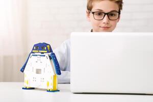 A young boy wearing glasses is sitting at a desk, using a laptop computer. He has a colorful, robotic toy on the desk in front of him. photo
