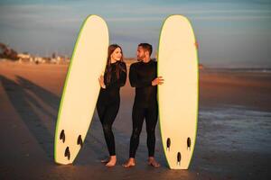 A young couple stands on a sandy beach at sunset, holding their surfboards. The sun is setting in the background, and the sky is a beautiful orange and pink. photo