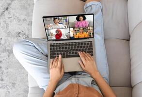 An overhead view of a person sitting on a couch, using a laptop to chat with four other people. The person is focused on the laptop screen, with their hands on the keyboard. photo