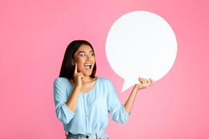 A young Asian woman with long, dark hair is standing in front of a pink background. She is holding a large, blank speech bubble in her right hand, and she is looking at it with a surprised expression photo