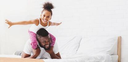 A little black girl with curly hair is riding on her fathers shoulders. Both are smiling and looking at the camera. They are on a white bed with white sheets and pillows, copy space photo