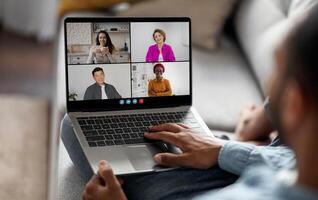 A man sits on a couch with a laptop in his lap. The screen displays a conference, showing four individuals in separate squares. The man is using his right hand to interact with the laptop photo
