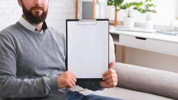 Confident psychotherapist showing white blank clipboard at camera, sitting in office, free space for announcement photo