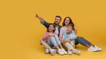 A joyful family is sitting closely with a father pointing at something out of frame, creating a moment of togetherness and curiosity, pointing at copy space on yellow background photo
