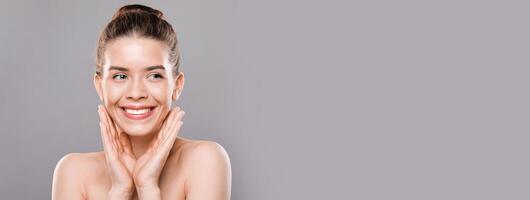Face care concept. Cheerful young woman touching her face and looking at copy space over gray studio background photo