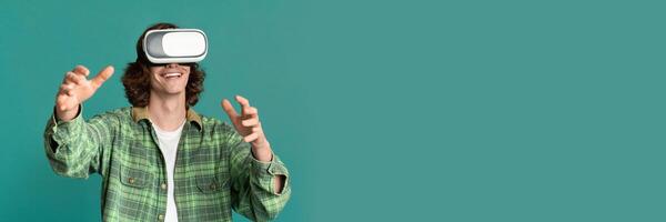Digital world fun. Young guy in virtual reality headset playing computer games on turquoise background, copy space, web-banner photo