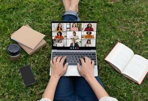 Lady is sitting on green grass, using a laptop for a call. There are multiple people on the screen, and the person is typing on the keyboard. photo