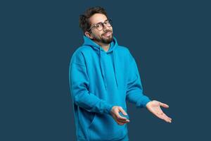 A young man with glasses is wearing a bright blue hoodie and displaying a confused expression. He raises his hands in a questioning gesture, suggesting uncertainty or disbelief photo