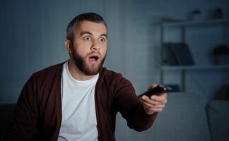 A man with a shocked expression is sitting on his couch at home in the evening, holding a TV remote and watching an unexpected scene on the screen. photo