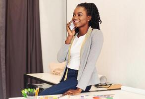 African American woman is seated at a desk, engrossed in a conversation on her cell phone. She appears focused and attentive as she communicates with the person on the other end of the line. photo