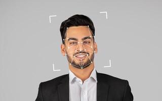 A young Indian man in a suit smiles confidently as a facial recognition overlay is displayed on his face in a studio setting. photo