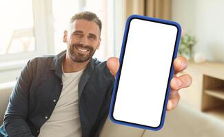 A man is seated on a couch while actively holding up a cell phone in his hand. He appears focused on the devices screen, engrossed in whatever content he is viewing. photo