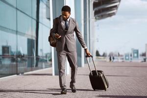 Black businessman is using his phone while standing with luggage outside a modern airport. The mood is busy and professional, capturing a moment of travel and communication. photo