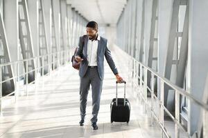 Black businessman in a suit checking his phone while walking by modern airport with a rolling suitcase, copy space photo