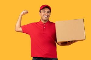A cheerful delivery worker man in a red cap and polo shirt holds a cardboard box while flexing his arm, showing enthusiasm and strength. photo