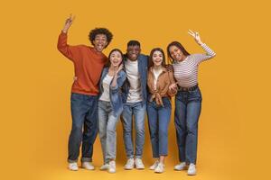 un grupo de cinco joven adultos estar juntos en frente de un brillante amarillo antecedentes. ellos son todas sonriente y reír, con su brazos alrededor cada otro. foto