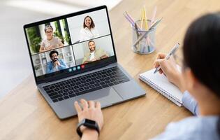 A woman trainee sits at a desk with a laptop open, participating in a conference meeting. The screen displays four people, each in a separate tile. photo