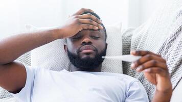 African American man with a beard lies in bed with a blanket covering him. He holds a thermometer in his hand and has his other hand on his forehead, looking down and appearing unwell. photo