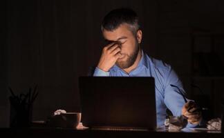 A tired and frustrated man rubs his eyes while working on a laptop late at night. The dark surroundings and crumpled papers suggest a challenging work environment, indicative of stress and fatigue. photo