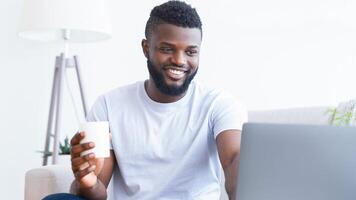 Black man is sitting on a couch in his home, smiling and using a laptop. He is holding a white mug in his right hand. The man is wearing a white t-shirt and appears to be enjoying his work. photo