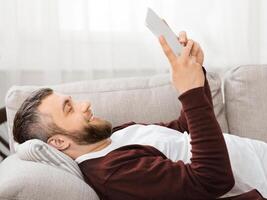 A bearded man is lying on a comfortable couch, using a tablet. The living room is well-lit with soft natural daylight coming in through the curtains, creating a serene and relaxing atmosphere. photo
