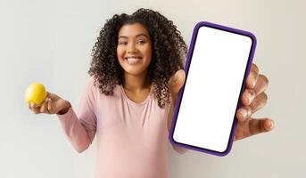 African American woman is holding a cell phone in one hand and an apple in the other hand. She appears to be looking at the screen of the phone, with the apple held casually. photo